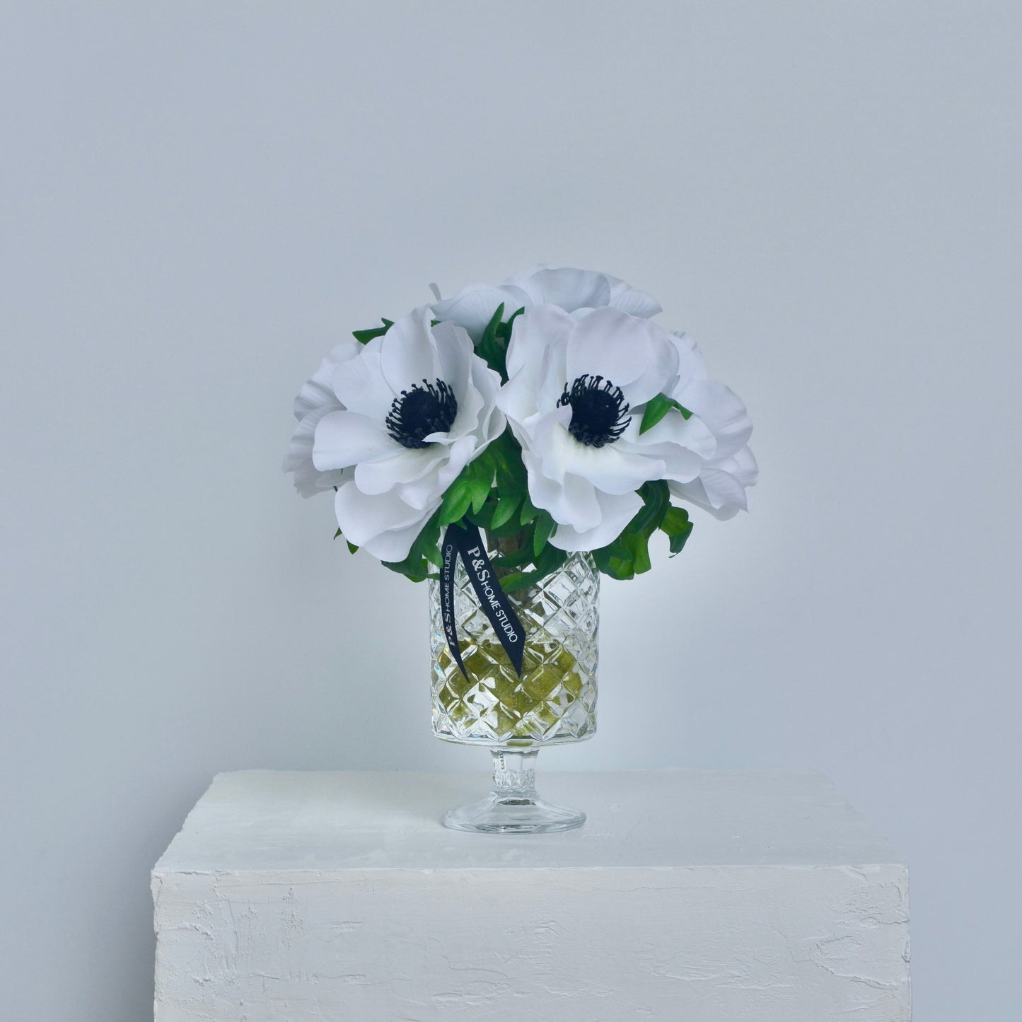 White flowers with green leaves in a glass vase on a white surface with a light gray background