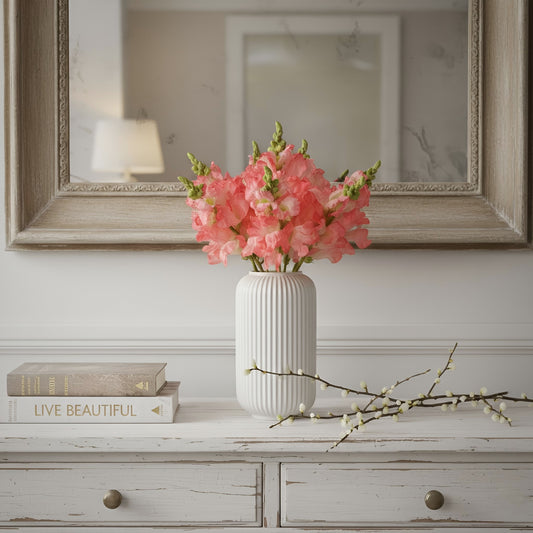 White vase with pink flowers on a wooden surface with books and a mirror in the background