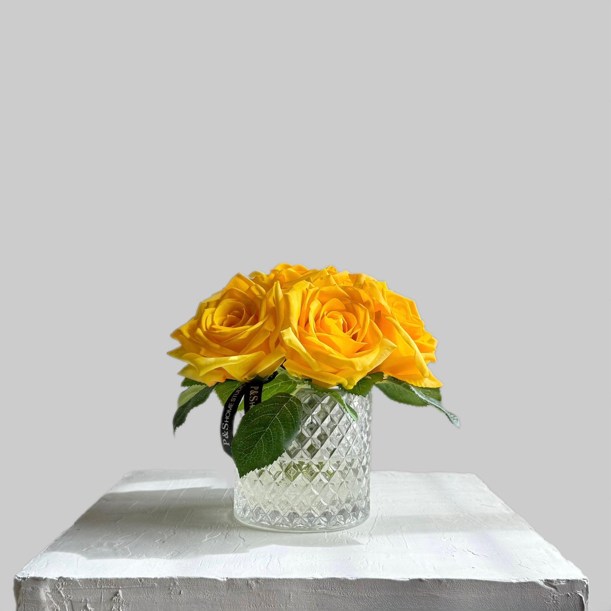 Clear glass vase with yellow roses on a white surface and light gray background