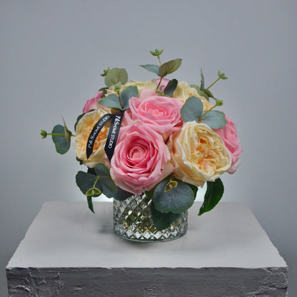 Bouquet of pink and yellow roses with greenery in a clear glass vase on a gray background